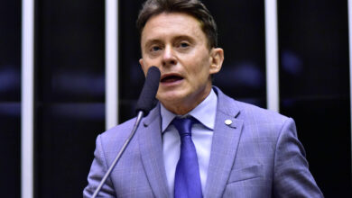 Um homem em um terno cinza claro e gravata azul está falando em um microfone em um ambiente de parlamento. Ele parece estar em uma posição de destaque, com um fundo que inclui painéis verticais. O homem tem cabelo castanho e está expressando-se de forma enérgica.