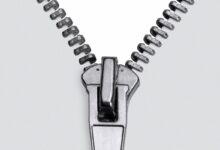 Zíper metálico prateado, detalhe em macro, com o cursor posicionado na parte inferior, mostrando os dentes se separando em um fundo branco e limpo. A imagem foca na textura e nos elementos mecânicos do fecho, transmitindo a ideia de abertura e funcionalidade.