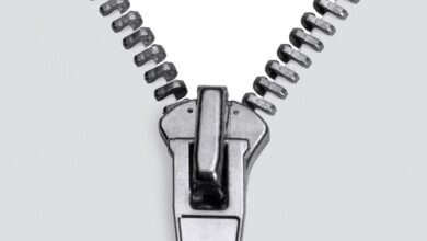Zíper metálico prateado, detalhe em macro, com o cursor posicionado na parte inferior, mostrando os dentes se separando em um fundo branco e limpo. A imagem foca na textura e nos elementos mecânicos do fecho, transmitindo a ideia de abertura e funcionalidade.