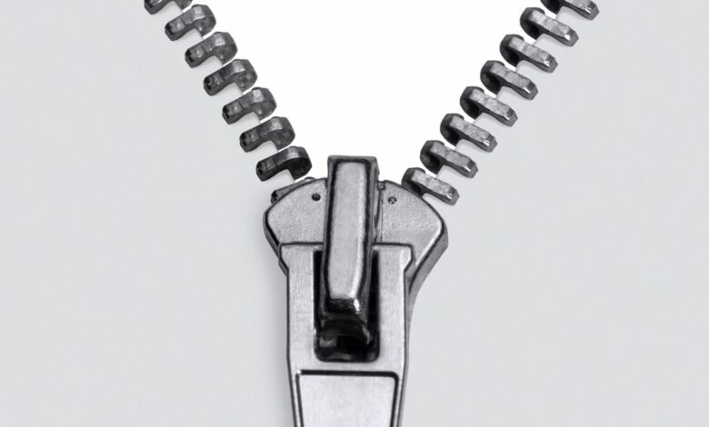 Zíper metálico prateado, detalhe em macro, com o cursor posicionado na parte inferior, mostrando os dentes se separando em um fundo branco e limpo. A imagem foca na textura e nos elementos mecânicos do fecho, transmitindo a ideia de abertura e funcionalidade.