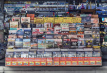 A imagem mostra uma banca de jornal com uma variedade de revistas e jornais expostos. A banca está organizada em prateleiras, com capas visíveis de diferentes publicações. À direita, uma mulher está de pé, usando uma máscara facial. O ambiente é iluminado e a banca é coberta por uma lona azul na parte superior.