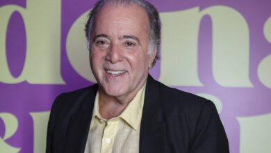 Um homem sorridente está posando para a foto em um evento. Ele usa um paletó preto e uma camisa amarela. O fundo é roxo com letras grandes em amarelo que dizem