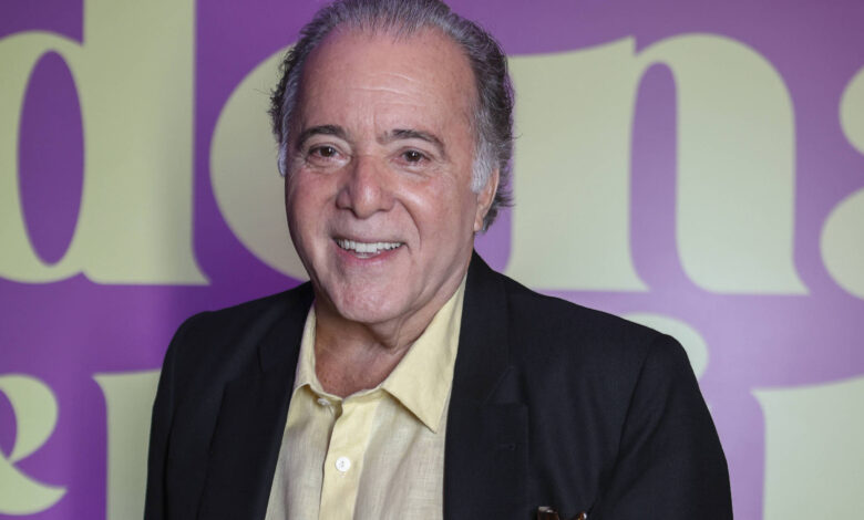 Um homem sorridente está posando para a foto em um evento. Ele usa um paletó preto e uma camisa amarela. O fundo é roxo com letras grandes em amarelo que dizem