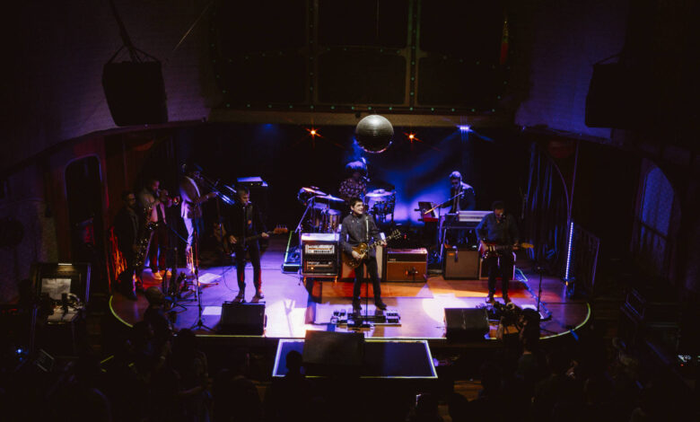 A imagem mostra uma banda se apresentando em um palco iluminado. Há vários músicos tocando instrumentos, incluindo guitarras e teclados. O palco é decorado com luzes coloridas e um grande disco de espelho pendurado no teto. A plateia é visível na parte inferior da imagem, com algumas pessoas assistindo ao show.
