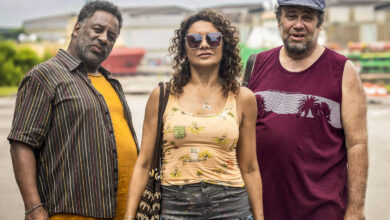 Três pessoas estão em pé em um ambiente urbano, possivelmente próximo a um porto. À esquerda, um homem de pele escura, com cabelo curto e barba, usa uma camisa listrada e calças escuras. No centro, uma mulher de pele clara, com cabelo cacheado e óculos escuros, veste uma blusa amarela com estampas florais e uma saia longa. À direita, um homem de pele clara, com cabelo curto e barba, usa uma camiseta roxa e calças brancas. Ao fundo, há estruturas industriais e equipamentos de porto.