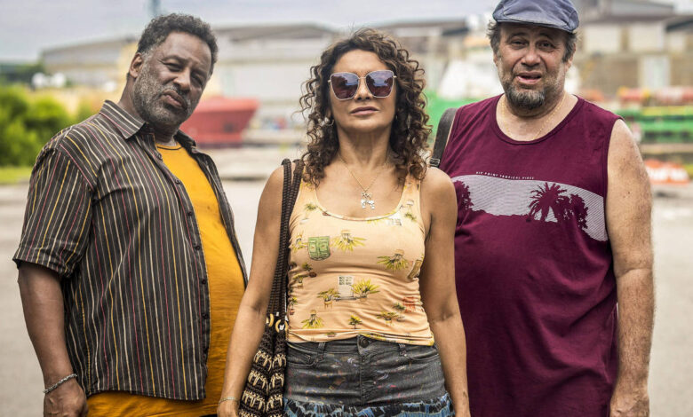 Três pessoas estão em pé em um ambiente urbano, possivelmente próximo a um porto. À esquerda, um homem de pele escura, com cabelo curto e barba, usa uma camisa listrada e calças escuras. No centro, uma mulher de pele clara, com cabelo cacheado e óculos escuros, veste uma blusa amarela com estampas florais e uma saia longa. À direita, um homem de pele clara, com cabelo curto e barba, usa uma camiseta roxa e calças brancas. Ao fundo, há estruturas industriais e equipamentos de porto.