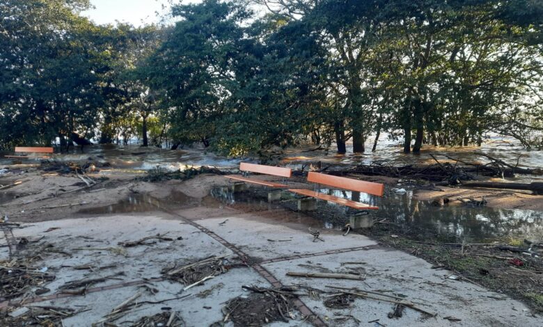 A imagem mostra uma área inundada, com água cobrindo parte do solo e detritos espalhados. Há bancos de madeira laranja visíveis, parcialmente submersos. Ao fundo, árvores estão presentes, e a luz do sol ilumina a cena, indicando que é durante o dia. A área parece ter sofrido os efeitos de uma enchente recente.