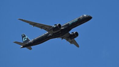 Um avião comercial está voando em um céu azul claro. O avião é de cor preta com detalhes em verde e possui a inscrição
