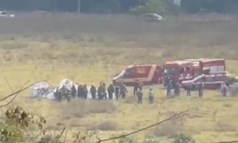 A imagem mostra um campo aberto onde ocorreu um acidente com uma aeronave. Há várias pessoas reunidas em torno do local do acidente, incluindo equipes de emergência. Ao fundo, é possível ver duas ambulâncias e um veículo de resgate. O ambiente é predominantemente seco e com vegetação baixa.