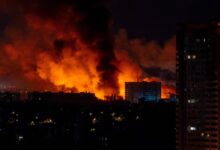 A imagem mostra um grande incêndio à noite, com chamas intensas e fumaça densa subindo em direção ao céu. No fundo, há edifícios iluminados pela luz do fogo, enquanto a parte inferior da imagem apresenta uma área urbana com várias luzes acesas. O céu está escuro, contrastando com o brilho laranja das chamas.