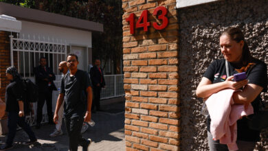 A imagem mostra a entrada de um edifício com um portão branco e um número 143 em destaque na parede de tijolos. Algumas pessoas estão saindo do local, vestindo roupas escuras. À direita, uma mulher está parada, segurando um celular e um objeto rosa, enquanto observa o movimento. Ao fundo, há outras pessoas e um segurança em terno.