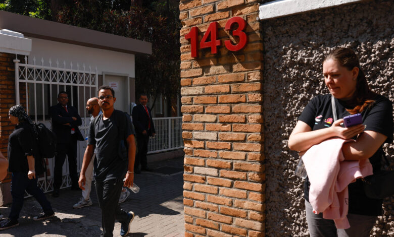 A imagem mostra a entrada de um edifício com um portão branco e um número 143 em destaque na parede de tijolos. Algumas pessoas estão saindo do local, vestindo roupas escuras. À direita, uma mulher está parada, segurando um celular e um objeto rosa, enquanto observa o movimento. Ao fundo, há outras pessoas e um segurança em terno.