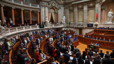 A imagem mostra o interior de um parlamento, com várias pessoas em pé e sentadas. O ambiente é grandioso, com colunas, estátuas e uma bandeira visível ao fundo. Há um painel digital que exibe um cronômetro. Os membros do parlamento estão vestidos formalmente, e a disposição das cadeiras é em formato semicircular.