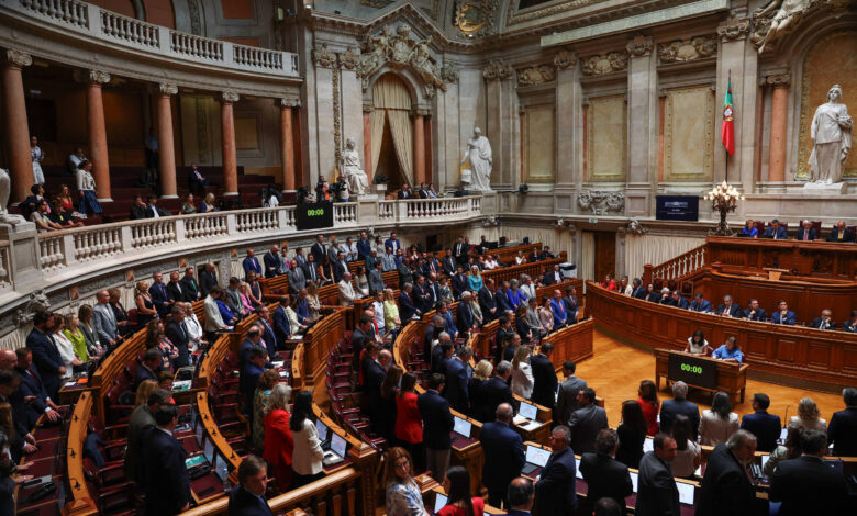 A imagem mostra o interior de um parlamento, com várias pessoas em pé e sentadas. O ambiente é grandioso, com colunas, estátuas e uma bandeira visível ao fundo. Há um painel digital que exibe um cronômetro. Os membros do parlamento estão vestidos formalmente, e a disposição das cadeiras é em formato semicircular.