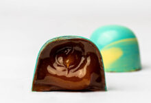 A imagem mostra um bombom de chocolate com uma casca colorida em azul e amarelo. O bombom está cortado ao meio, revelando um recheio de chocolate escuro e brilhante. Ao fundo, há outro bombom inteiro, também com a casca colorida, mas não cortado.