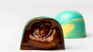 A imagem mostra um bombom de chocolate com uma casca colorida em azul e amarelo. O bombom está cortado ao meio, revelando um recheio de chocolate escuro e brilhante. Ao fundo, há outro bombom inteiro, também com a casca colorida, mas não cortado.