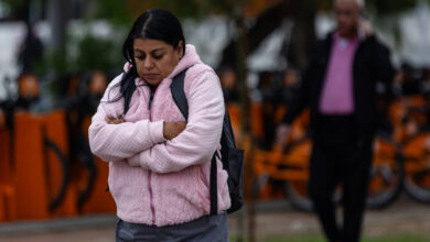 A imagem mostra uma mulher com cabelo longo e escuro, vestindo um colete rosa e calças cinzas, com os braços cruzados em frente ao corpo, aparentando estar pensativa ou triste. Ao fundo, há um homem com cabelo grisalho, usando uma camisa rosa e um casaco escuro, e bicicletas laranjas estacionadas. O ambiente parece ser ao ar livre, possivelmente em um parque, em um dia nublado.