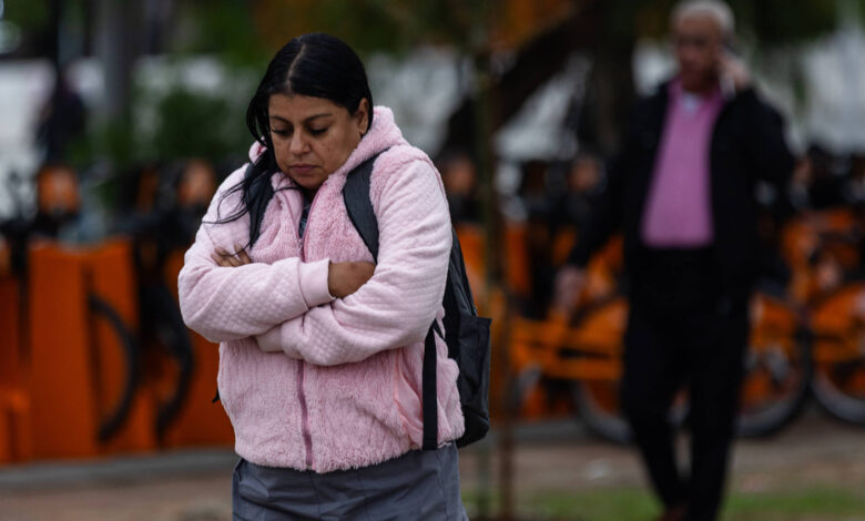 A imagem mostra uma mulher com cabelo longo e escuro, vestindo um colete rosa e calças cinzas, com os braços cruzados em frente ao corpo, aparentando estar pensativa ou triste. Ao fundo, há um homem com cabelo grisalho, usando uma camisa rosa e um casaco escuro, e bicicletas laranjas estacionadas. O ambiente parece ser ao ar livre, possivelmente em um parque, em um dia nublado.
