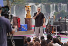 Um apresentador está em um palco ao ar livre, falando para uma plateia. Ao fundo, vê-se as Cataratas do Iguaçu. O apresentador usa uma camiseta escura e calças claras, segurando um microfone. À esquerda, há um boneco de um animal com aparência de onça, e ao lado, instrumentos musicais como guitarras estão dispostos. A plateia é composta por várias pessoas que assistem ao show.