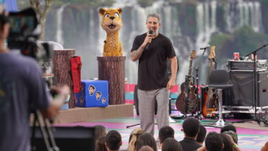 Um apresentador está em um palco ao ar livre, falando para uma plateia. Ao fundo, vê-se as Cataratas do Iguaçu. O apresentador usa uma camiseta escura e calças claras, segurando um microfone. À esquerda, há um boneco de um animal com aparência de onça, e ao lado, instrumentos musicais como guitarras estão dispostos. A plateia é composta por várias pessoas que assistem ao show.