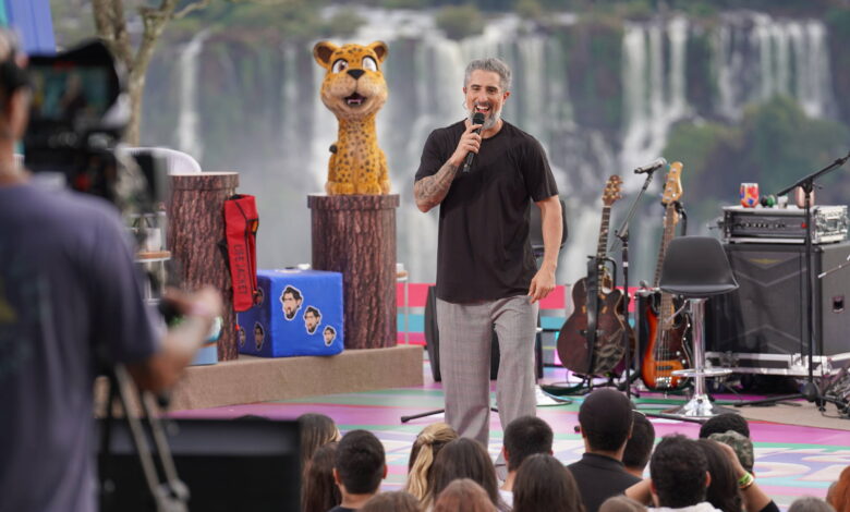 Um apresentador está em um palco ao ar livre, falando para uma plateia. Ao fundo, vê-se as Cataratas do Iguaçu. O apresentador usa uma camiseta escura e calças claras, segurando um microfone. À esquerda, há um boneco de um animal com aparência de onça, e ao lado, instrumentos musicais como guitarras estão dispostos. A plateia é composta por várias pessoas que assistem ao show.