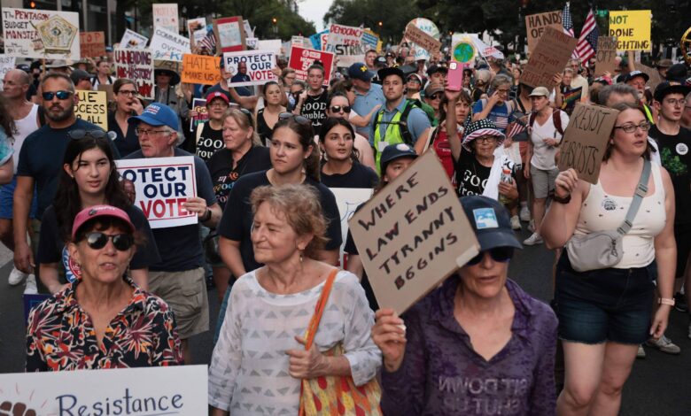 A imagem mostra uma grande multidão participando de uma manifestação. As pessoas estão segurando cartazes com mensagens como