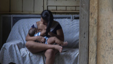 A imagem mostra uma mulher sentada em um sofá, amamentando um bebê. Ela está vestida com uma blusa listrada e calças. Ao fundo, uma outra mulher está de pé, olhando para a cena através de um espelho. A parede é de madeira e o ambiente parece ser uma casa simples, com janelas que mostram vegetação do lado de fora.
