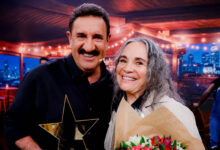 Na imagem, um homem e uma mulher estão sorrindo juntos em um ambiente iluminado. O homem, com bigode e cabelo curto, segura um troféu em forma de estrela. A mulher, com cabelo longo e grisalho, está segurando um buquê de flores coloridas. Ao fundo, há uma decoração com luzes e uma vista da cidade.