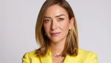 Retrato de uma mulher de cabelo loiro, usando blazer amarelo e joias douradas, com expressão confiante em fundo claro.