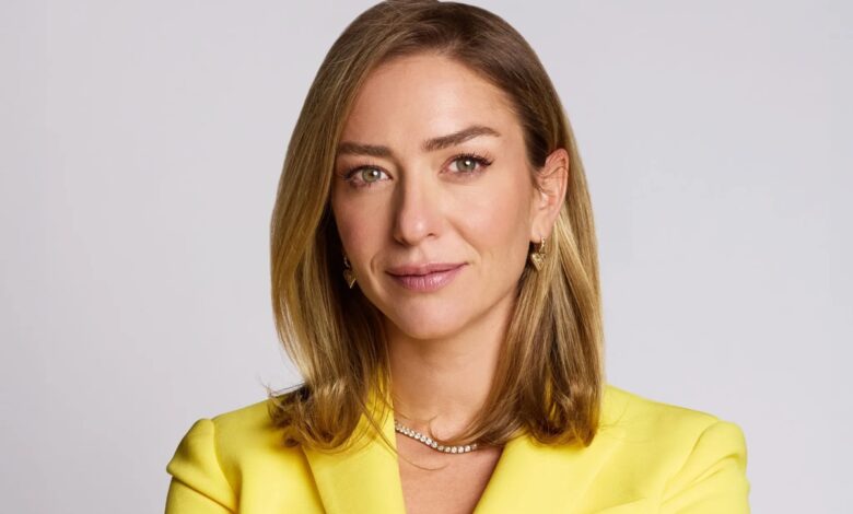 Retrato de uma mulher de cabelo loiro, usando blazer amarelo e joias douradas, com expressão confiante em fundo claro.