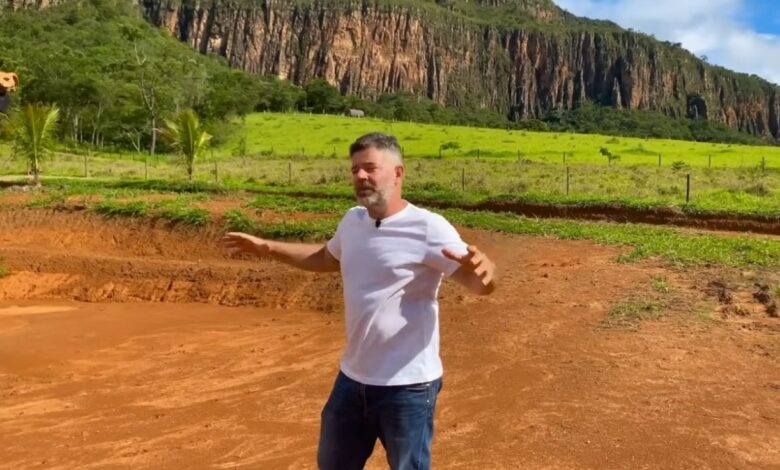 Homem em pé sobre solo vermelho em fazenda em Minas Gerais com paredão de pedra e vegetação ao fundo, em área de construção sustentável