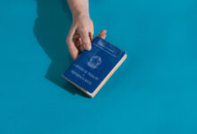 A imagem mostra uma mão segurando uma carteira de trabalho, que é de cor azul. A carteira está posicionada sobre uma superfície de cor azul clara. O texto na capa da carteira diz