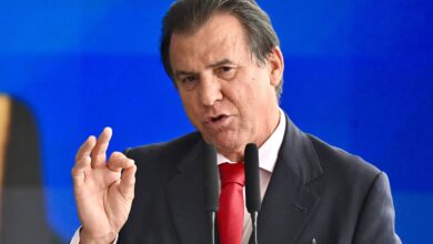 Um homem está falando em um microfone, gesticulando com a mão direita em um sinal de aprovação. Ele usa um terno escuro e uma gravata vermelha. O fundo é azul, sugerindo um ambiente formal.