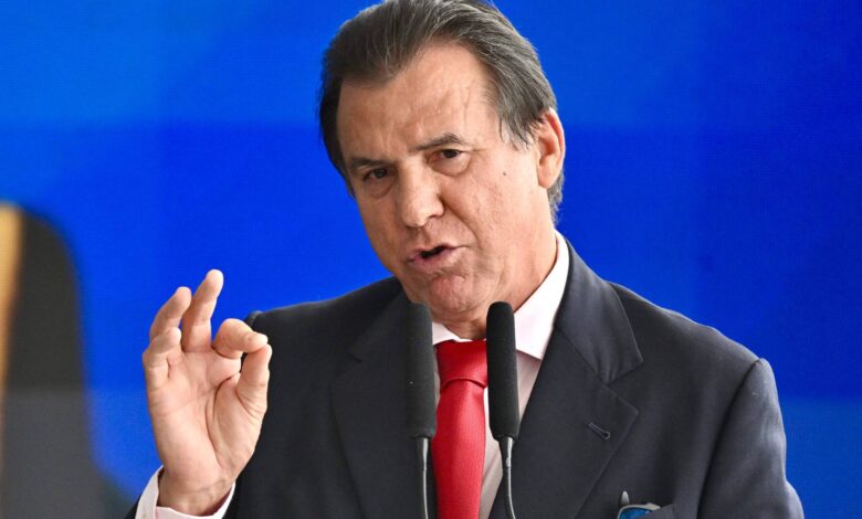 Um homem está falando em um microfone, gesticulando com a mão direita em um sinal de aprovação. Ele usa um terno escuro e uma gravata vermelha. O fundo é azul, sugerindo um ambiente formal.