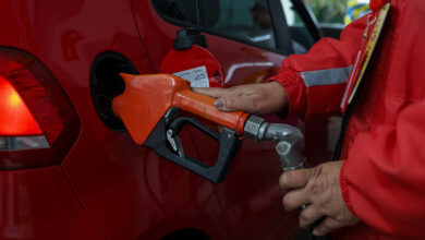 A imagem mostra um carro vermelho sendo abastecido com combustível. Uma pessoa, vestindo um uniforme vermelho, segura uma mangueira de abastecimento laranja, que está inserida no tanque do carro. O fundo é desfocado, mas é possível ver outros veículos e uma área de abastecimento.