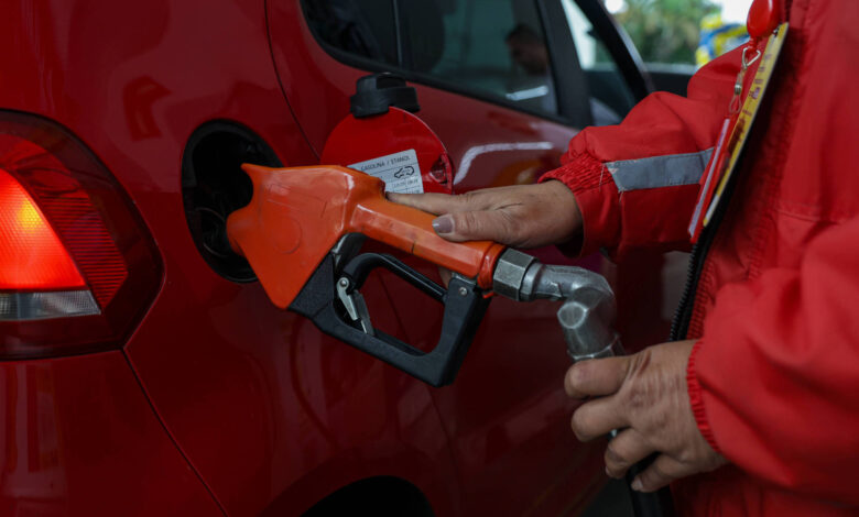 A imagem mostra um carro vermelho sendo abastecido com combustível. Uma pessoa, vestindo um uniforme vermelho, segura uma mangueira de abastecimento laranja, que está inserida no tanque do carro. O fundo é desfocado, mas é possível ver outros veículos e uma área de abastecimento.