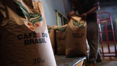 A imagem mostra sacos de café empilhados em um ambiente interno. Os sacos são de papel marrom e têm a inscrição