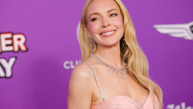 Uma mulher sorridente está em um evento de tapete vermelho, usando um vestido rosa claro com detalhes brilhantes. Ela tem cabelos longos e ondulados, e está usando joias. O fundo é roxo com texto que diz
