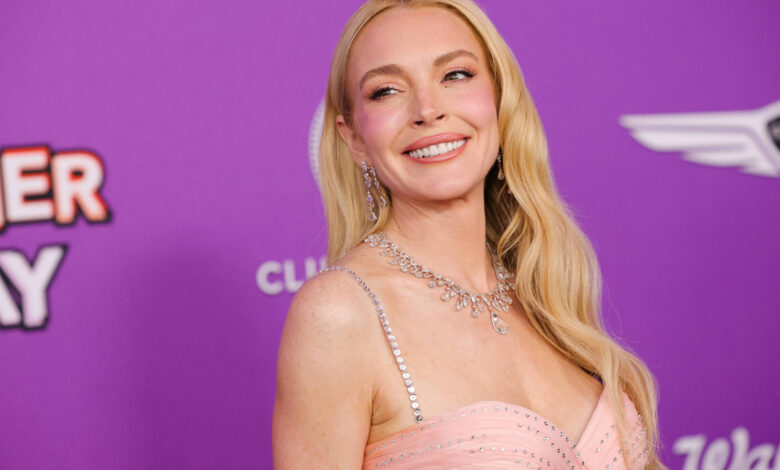 Uma mulher sorridente está em um evento de tapete vermelho, usando um vestido rosa claro com detalhes brilhantes. Ela tem cabelos longos e ondulados, e está usando joias. O fundo é roxo com texto que diz