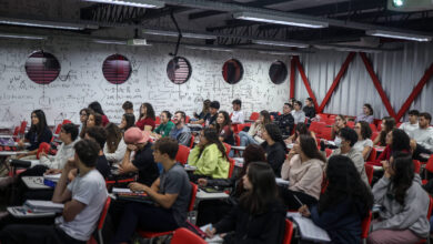 A imagem mostra uma sala de aula com muitos alunos sentados em cadeiras vermelhas. O ambiente é iluminado e as paredes estão cobertas com anotações e fórmulas matemáticas. Os alunos estão atentos, alguns olhando para frente e outros conversando. Há janelas redondas na parede, que permitem a entrada de luz natural.