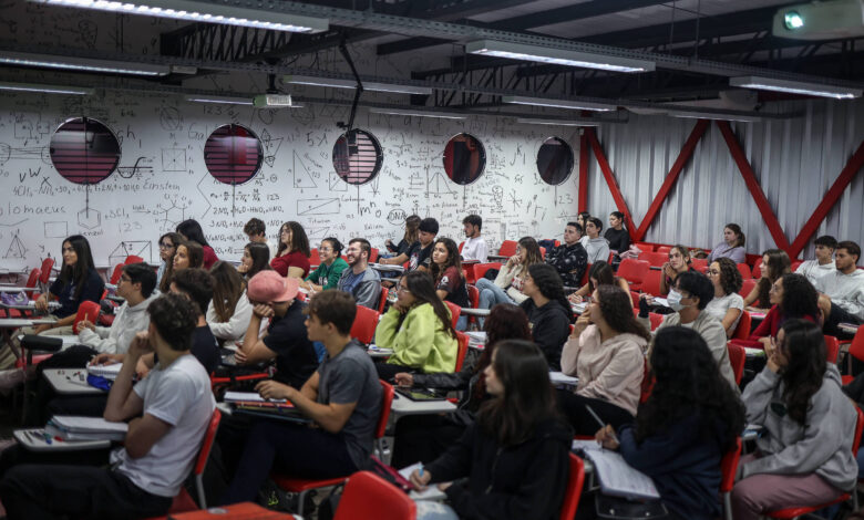 A imagem mostra uma sala de aula com muitos alunos sentados em cadeiras vermelhas. O ambiente é iluminado e as paredes estão cobertas com anotações e fórmulas matemáticas. Os alunos estão atentos, alguns olhando para frente e outros conversando. Há janelas redondas na parede, que permitem a entrada de luz natural.