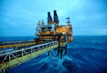 A imagem mostra uma plataforma de petróleo localizada no mar, com estruturas metálicas e chaminés visíveis. O céu está nublado e a água do mar apresenta ondas, refletindo uma tonalidade azul. A plataforma está iluminada, destacando-se contra o fundo escuro do céu e do mar.