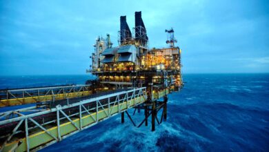 A imagem mostra uma plataforma de petróleo localizada no mar, com estruturas metálicas e chaminés visíveis. O céu está nublado e a água do mar apresenta ondas, refletindo uma tonalidade azul. A plataforma está iluminada, destacando-se contra o fundo escuro do céu e do mar.