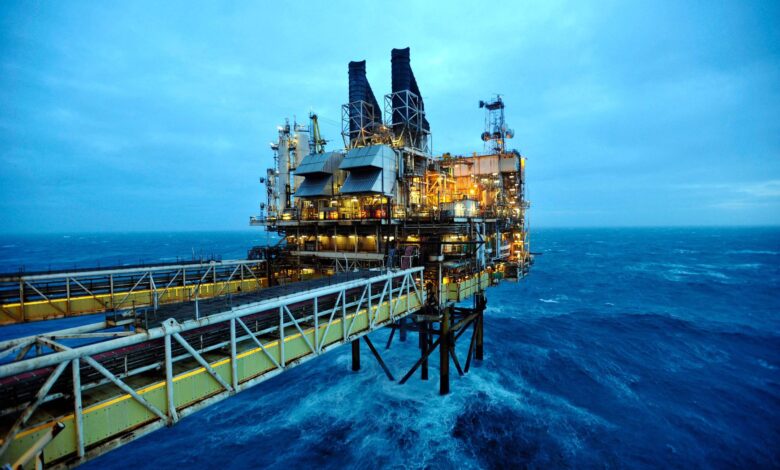 A imagem mostra uma plataforma de petróleo localizada no mar, com estruturas metálicas e chaminés visíveis. O céu está nublado e a água do mar apresenta ondas, refletindo uma tonalidade azul. A plataforma está iluminada, destacando-se contra o fundo escuro do céu e do mar.