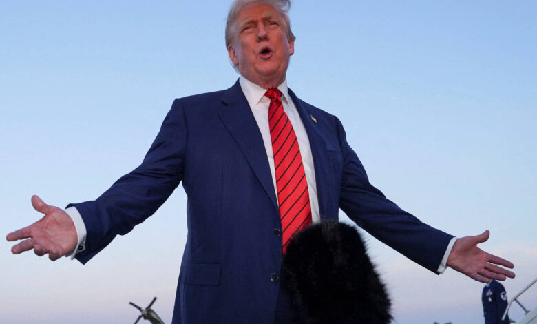 Imagem mostra Donald Trump, um homem em traje formal, com um terno azul e uma gravata vermelha, está de pé em uma pista de pouso, com os braços abertos e falando. Ele parece estar em uma entrevista ou discurso. Ao fundo, há uma pista de avião e algumas pessoas em trajes formais. Microfones estão posicionados próximos a ele, e o céu está claro.