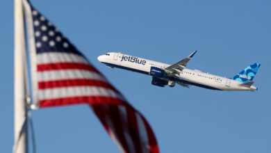 A imagem mostra um avião da companhia aérea JetBlue voando em direção ao céu, com a bandeira dos Estados Unidos em primeiro plano. O avião é branco com detalhes em azul e verde, enquanto a bandeira apresenta listras vermelhas e brancas e um campo azul com estrelas brancas.