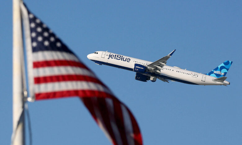 A imagem mostra um avião da companhia aérea JetBlue voando em direção ao céu, com a bandeira dos Estados Unidos em primeiro plano. O avião é branco com detalhes em azul e verde, enquanto a bandeira apresenta listras vermelhas e brancas e um campo azul com estrelas brancas.