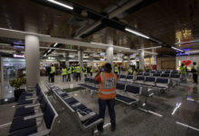 A imagem mostra o interior de um aeroporto, com várias cadeiras de espera em primeiro plano. Há pessoas vestindo coletes amarelos, algumas delas em grupos, e outras sozinhas. O ambiente é moderno, com iluminação artificial e lojas visíveis ao fundo. O piso é de granito e há plantas decorativas em algumas áreas.
