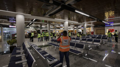 A imagem mostra o interior de um aeroporto, com várias cadeiras de espera em primeiro plano. Há pessoas vestindo coletes amarelos, algumas delas em grupos, e outras sozinhas. O ambiente é moderno, com iluminação artificial e lojas visíveis ao fundo. O piso é de granito e há plantas decorativas em algumas áreas.