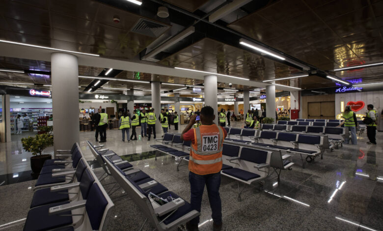 A imagem mostra o interior de um aeroporto, com várias cadeiras de espera em primeiro plano. Há pessoas vestindo coletes amarelos, algumas delas em grupos, e outras sozinhas. O ambiente é moderno, com iluminação artificial e lojas visíveis ao fundo. O piso é de granito e há plantas decorativas em algumas áreas.